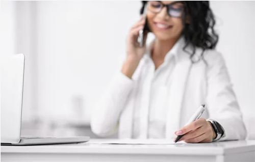 Woman taking notes while on the phone in front of a laptop