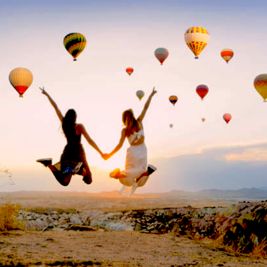 Two women floating, holding hands, and surrounded by hot air balloons.